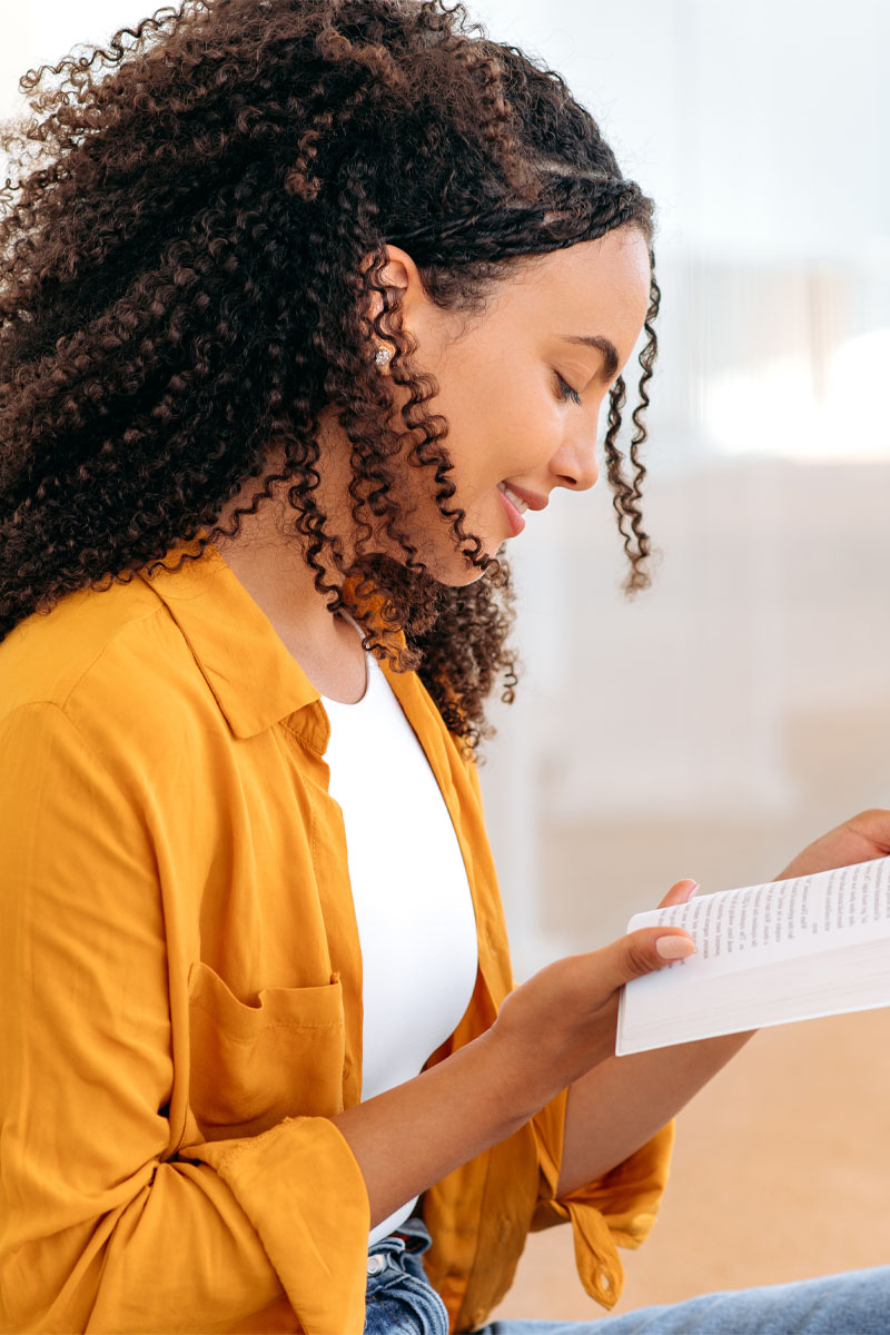 Side view photo of a lovely stylish curly haired brazilian or hispanic female student, sit near the university campus, focused reading her favorite a book